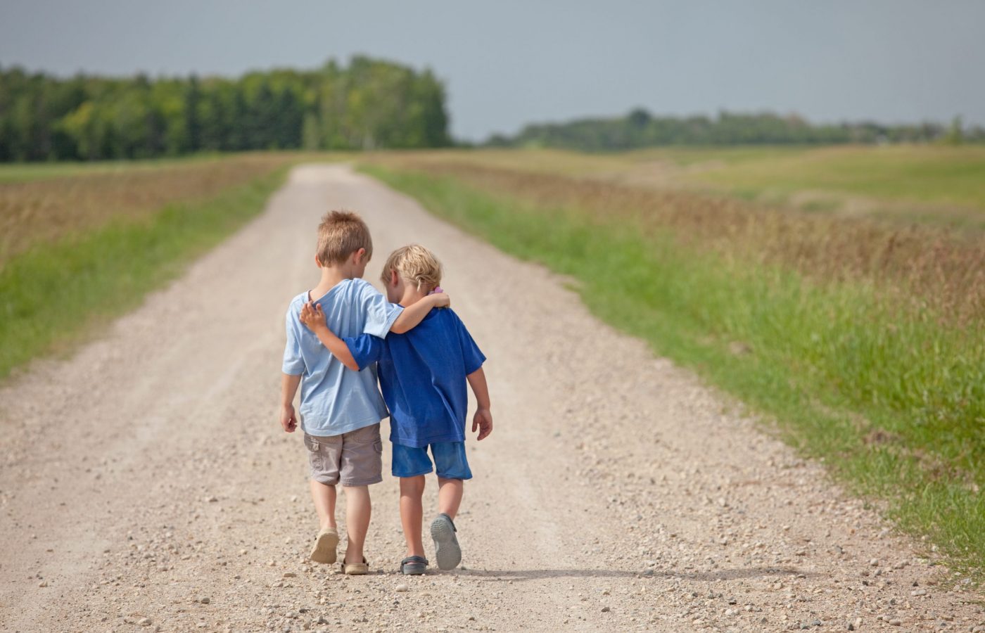 Two young boys walking arm in arm down a rural country gravel road. Prairie scene. Elementary aged caucasian children. Horizontal color image. Additional themes include friendship, bonding, boys, embracing, hugging, love, relationships, brothers, kindergarten, preschool, talking, care, best friends, and summer.